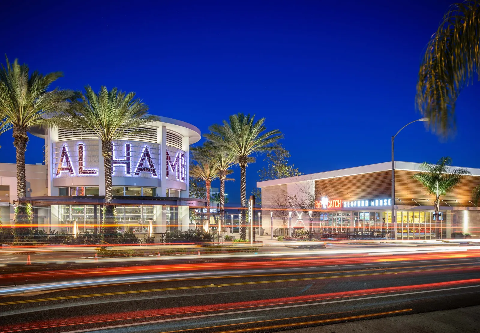 Alhambra Place at night: palm trees and a lit round tower with the Alhambra sign, shops and restaurants along the street, and red and white light trails from passing cars under a deep blue sky.
